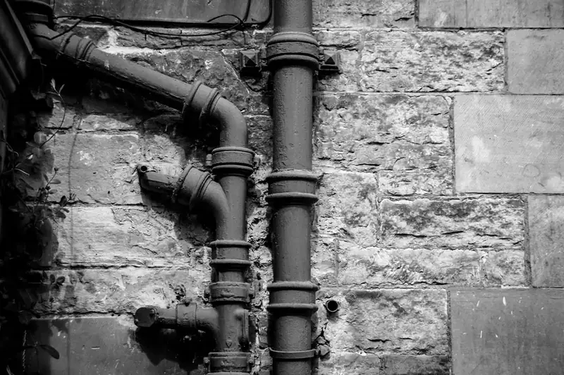 Black and white photo of old cast iron downpipes with offset bends fixed to a stone wall