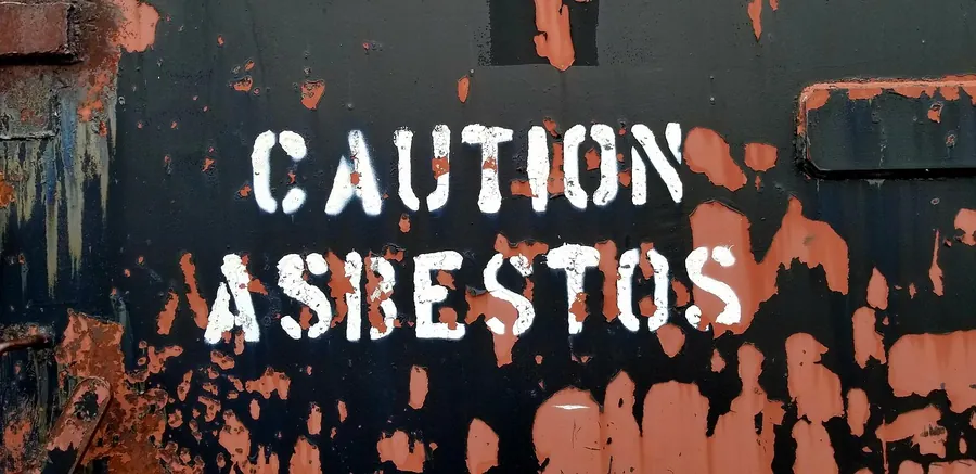 Caution asbestos warning stencilled in white on a dark, rust-streaked metal panel