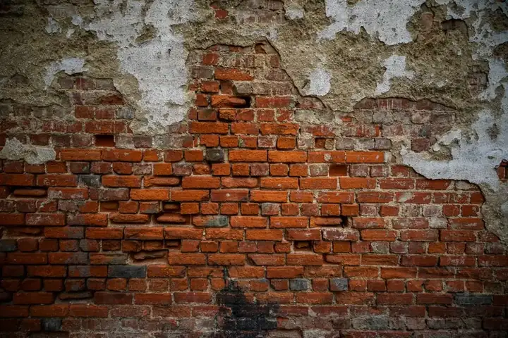 Patchy damp plaster flaking away from an old red brick wall, exposing the brickwork underneath