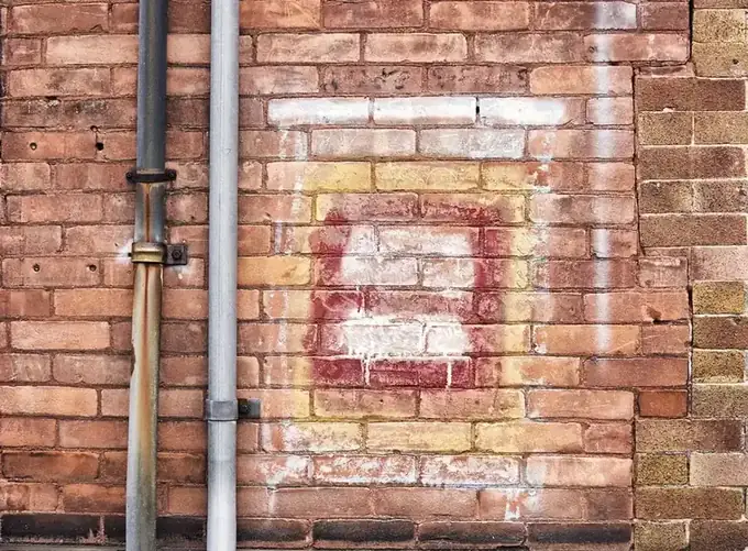 Two downpipes, one metal and one grey plastic, running down an old brick wall with a faded painted square
