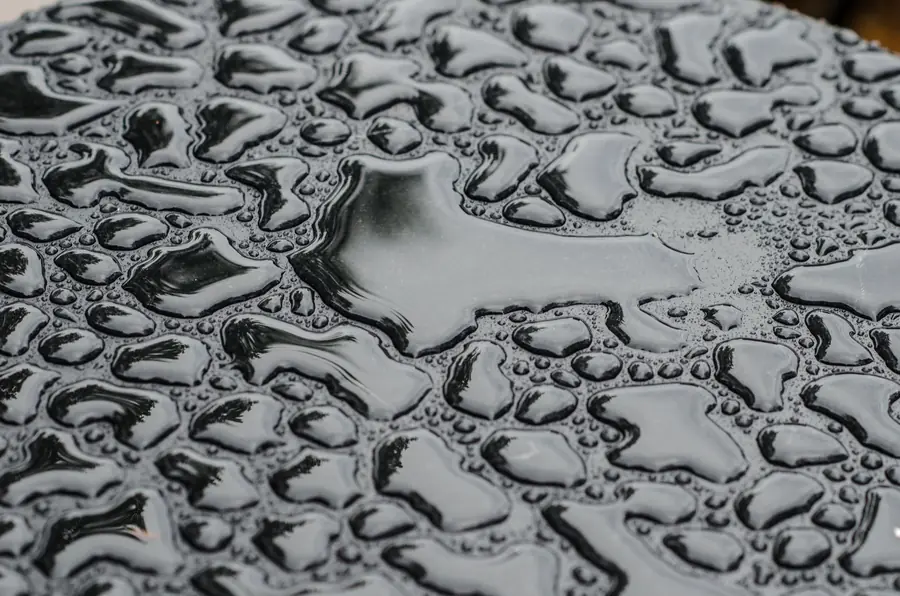 Rainwater beading and pooling on a smooth dark surface, similar to water sitting on a flat roof membrane