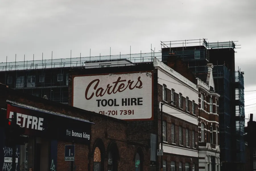 Scaffolding rising above brick buildings and an old tool hire sign on a south-east London street, the access cost behind many roof repairs