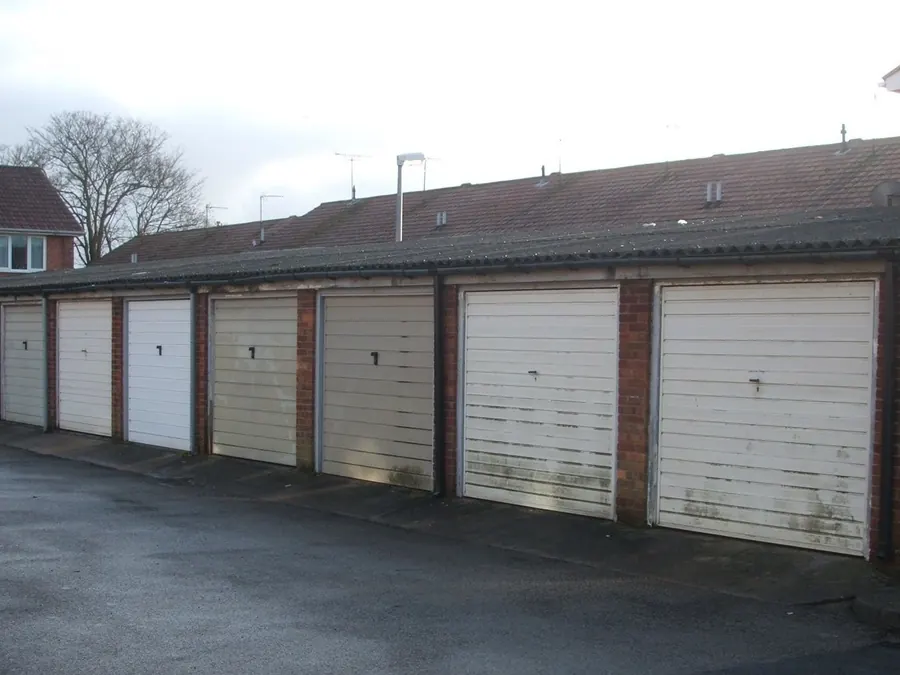 Brick garage block with white doors under a long corrugated sheet roof, backed by terraced houses