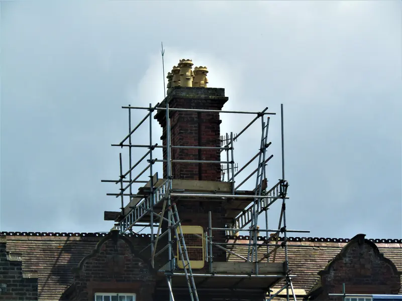 Tall brick chimney stack with clay pots, wrapped in a scaffold platform above a tiled roof