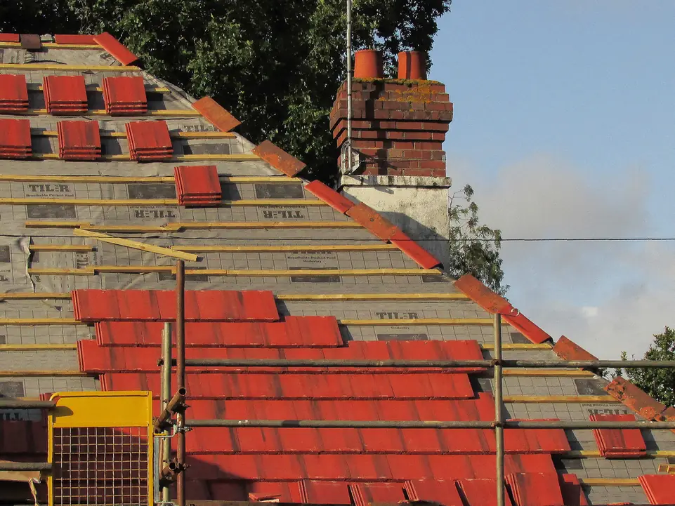 New red roof tiles being fixed onto fresh battens and breathable membrane during a full re-roof, with a brick chimney stack at the ridge