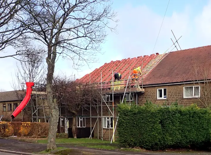 Roofers on scaffold re-tiling the roof of a pair of semi-detached houses, with a rubble chute down to a skip