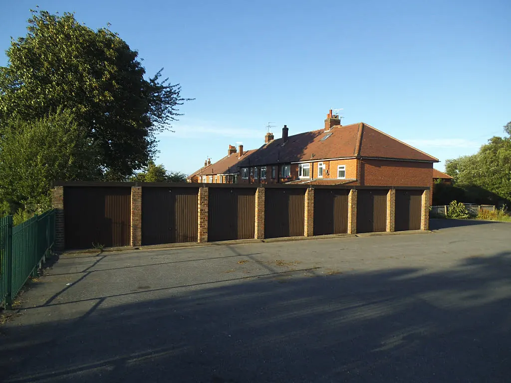 Row of flat-roofed brick lock-up garages with brown up-and-over doors in front of red brick semi-detached houses